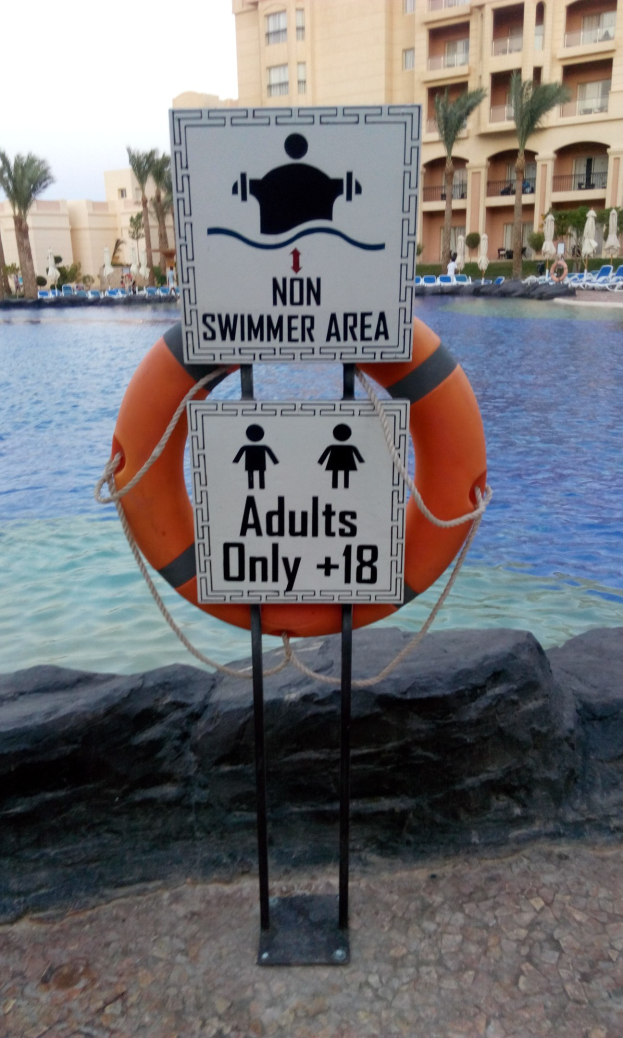 Ein Schwimmbecken mit einem "Non Swimmer Area Adults Only"-Schild im Vordergrund, umgeben von Bäumen, Gebäuden und einem klaren blauen Himmel, mit einem Rettungsring in der Mitte des Pools.