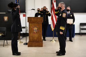 Gruppe uniformierter Personen in Masken auf einem Podium während einer Vereidigung für einen neuen Feuerchef, der eine Urkunde hält, mit Flaggen, Stühlen, Tischen und einer Wand im Hintergrund.