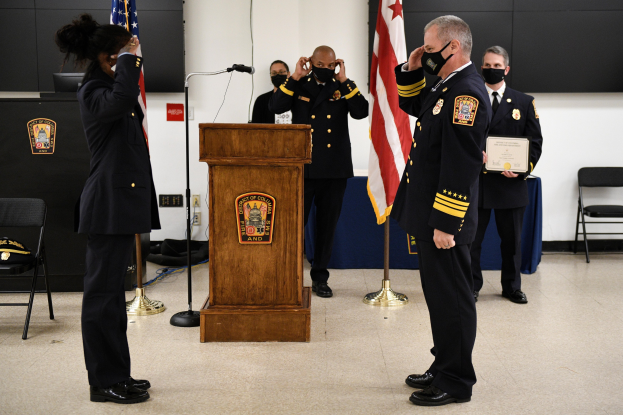 Gruppe uniformierter Personen in Masken auf einem Podium während einer Vereidigung für einen neuen Feuerchef, der eine Urkunde hält, mit Flaggen, Stühlen, Tischen und einer Wand im Hintergrund.