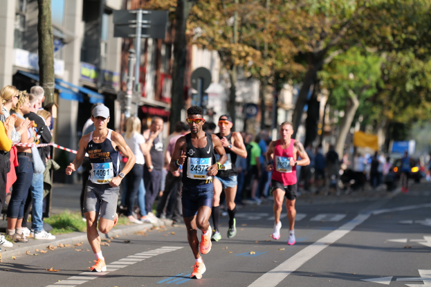 Läufer bei einem Marathon auf einer Stadtstraße, mit Zuschauern auf der linken Seite, Bäumen, Gebäuden und einem klaren blauen Himmel im Hintergrund; alle Läufer tragen Turnschuhe und Hüte.