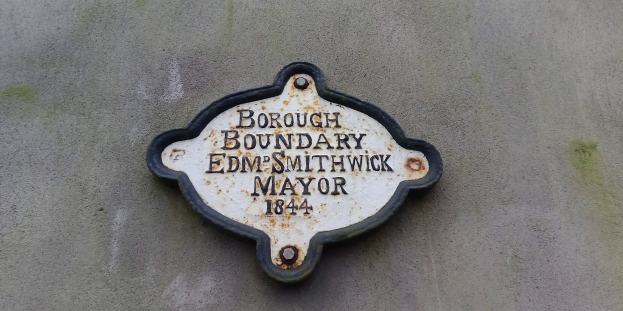 Metallplatte an einer Gebäudeseite, die die Borough-Grenze markiert, mit "Borough Boundary Edm Smithwick Mayor" in fetten weißen Buchstaben auf einem schwarzen Hintergrund.