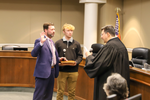 Ein Mann in Anzug und Krawatte steht neben einem Richter in einer Robe in einem Gerichtssaal, mit Stühlen, Tischen, einer Fahne, einer Säule und einer sitzenden Person im Hintergrund.