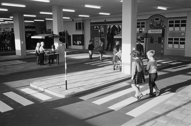 Schwarzes und weißes Foto von Kindern, die eine Straße an einer Ampel überqueren, mit Fußgängern in der Nähe, einer Verkehrsampel und strukturellen Säulen mit Deckenleuchten im Hintergrund.