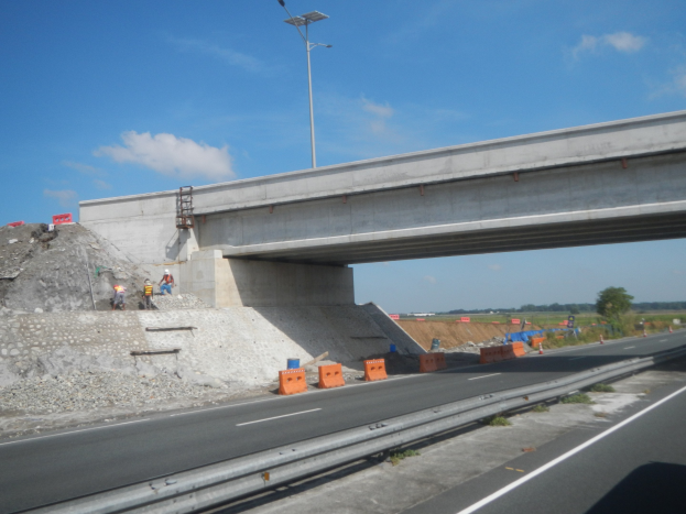 Autobahn mit einer Brücke darüber, ein darunter fahrendes Auto und Bauarbeiter am Straßenrand, umgeben von Verkehrskegeln, Steinen und einem Lichtmast, mit Bäumen und einem bewölkten Himmel im Hintergrund.