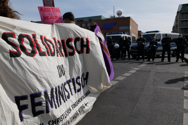 Gruppe von Menschen, die mit einem 'Solidarität und Feminismus' -Schild die Straße entlangmarschieren, mit parkenden Fahrzeugen, Gebäuden, einer Parabolantenne und einem klaren blauen Himmel im Hintergrund.