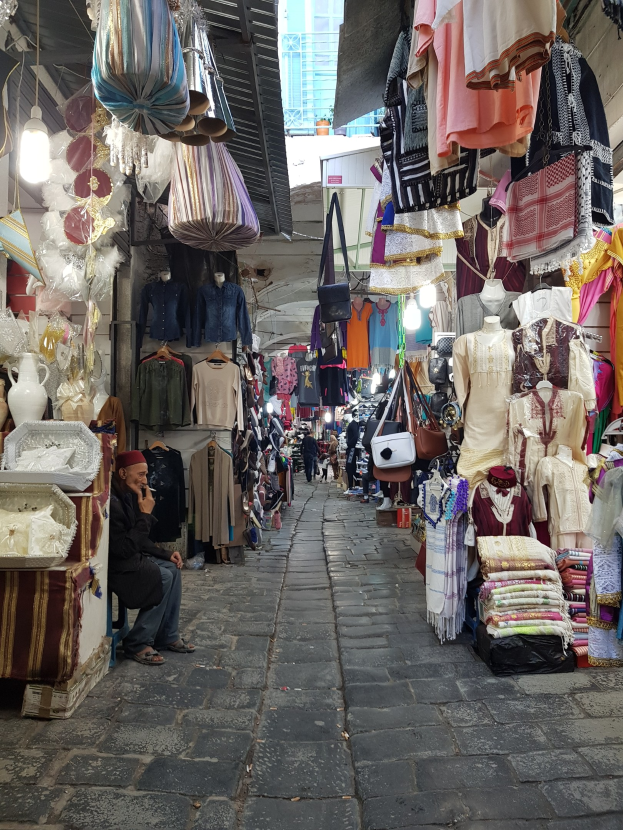 Ein belebter Straßenmarkt in der Altstadt von Jerusalem mit Ständen, die Kleidung, Taschen, Lampen und andere Artikel verkaufen, umgeben von Menschen, die durch die Gassen schlendern und ein Gebäude im Hintergrund.