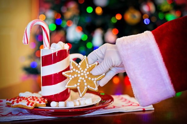 Eine Person in einem Weihnachtsmannhut hält einen Becher heiße Schokolade und einen Zuckerstange in der Hand und hat einen Teller mit Keksen und ein Tuch auf dem Tisch vor sich, vor einem Weihnachtsbaum und einer Wand.
