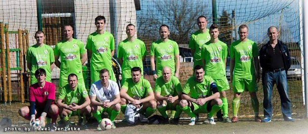 Eine Gruppe von Männern in grünen T-Shirts steht auf einem Fücballfeld mit einem Ball und einem Tor, einige hocken, mit Bäumen, Gebäuden und Fahrzeugen im Hintergrund.