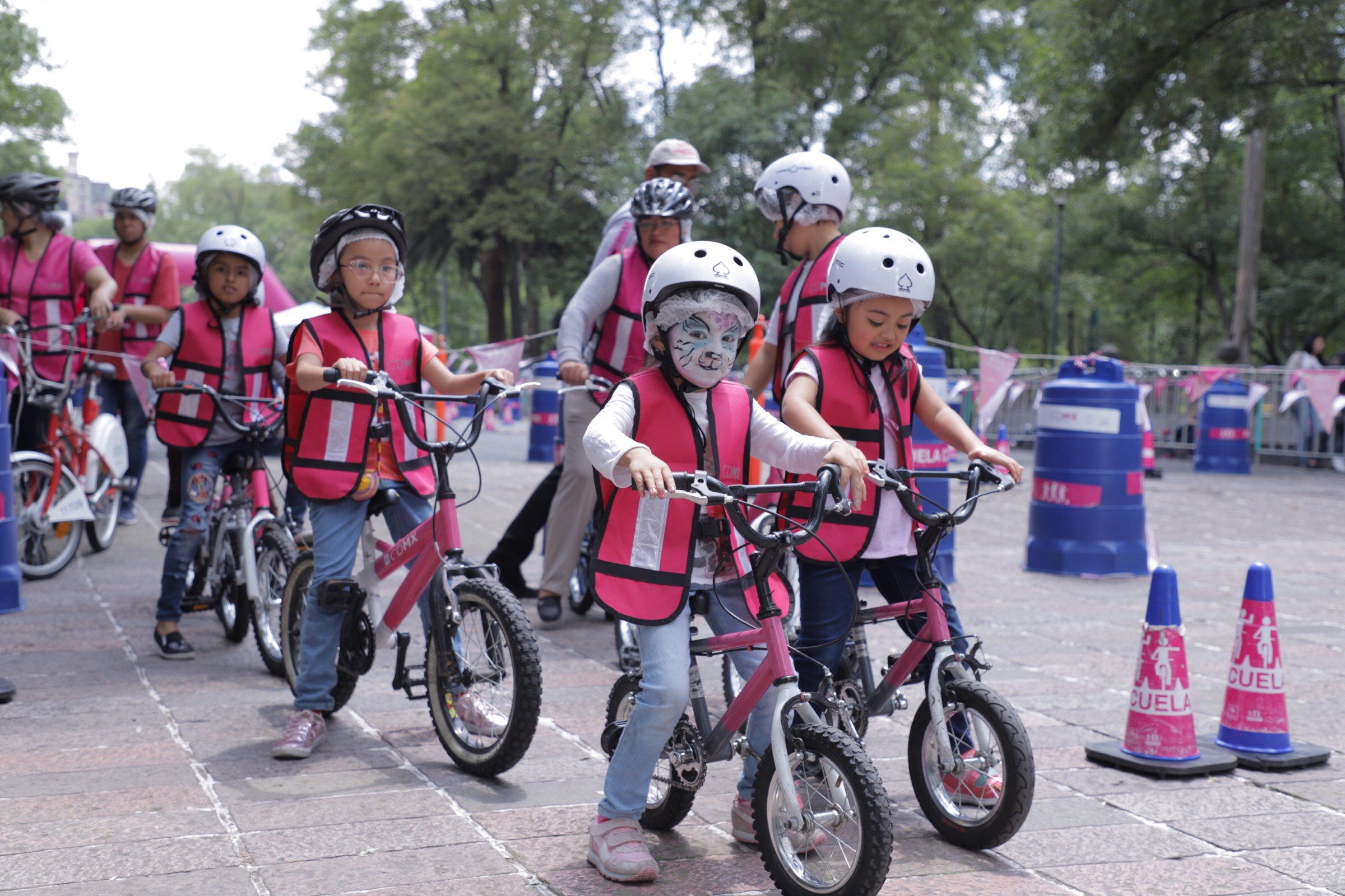 Kinder mit Helmen fahren auf einer Straße mit Verkehrskegeln Fahrrad, einige mit Gesichtsbemalung, mit Bäumen und einem klaren blauen Himmel im Hintergrund.