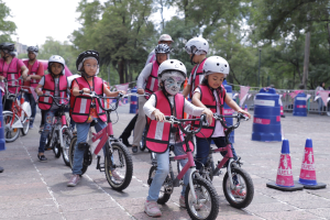 Kinder mit Helmen fahren auf einer Straße mit Verkehrskegeln Fahrrad, einige mit Gesichtsbemalung, mit Bäumen und einem klaren blauen Himmel im Hintergrund.