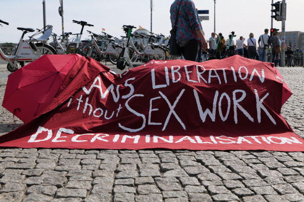 Gruppe von Menschen, die auf einer Stadtstraße protestieren und Regenschirme halten sowie ein rotes Banner mit der Aufschrift 'Befreiung ohne Entkriminalisierung der Sexarbeit' schwenken, mit Fahrrädern und städtischer Infrastruktur im Hintergrund.