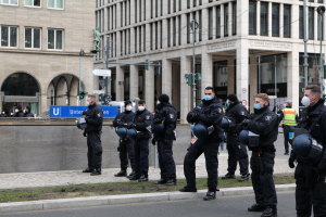 Eine Gruppe von Polizisten in schwarzen Uniformen und Masken steht vor einem Gebäude mit Glasfenstern und Säulen in Berlin, Deutschland, einige halten Helme, mit Laternenmasten, Verkehrsampeln, Schildern mit Text, einer Statue, anderen Menschen und einer grasbewachsenen Fläche im Hintergrund.