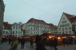 Ein belebter Weihnachtsmarkt in Tallinn, Estland mit Menschen, Ständen, beleuchteten Gebäuden, Bäumen und einem klaren blauen Himmel.
