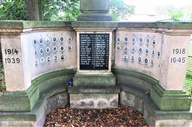 Ein Holocaust-Gedenkmonument in einem jüdischen Friedhof in Berlin, mit einer Text- und Nummerntafel an seiner Wand, umgeben von Bäumen, einem Zaun und verstreuten trockenen Blättern.