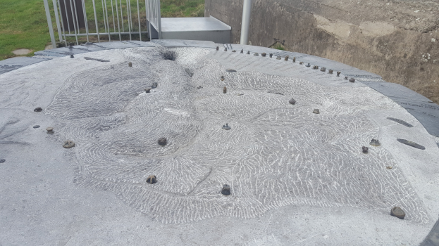 Große Betonstruktur mit kreisförmigem Design oben drauf, umgeben von einem Metallzaun, einem Pfahl, einer Wand und Grasboden.