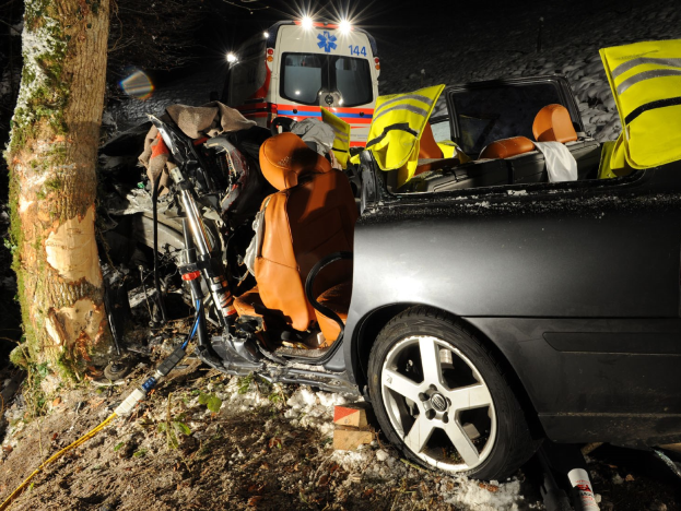 Ein geschädigtes Auto am Straßenrand mit einem Rettungswagen im Hintergrund, umgeben von Bäumen.