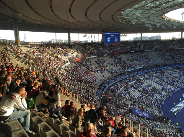 Große Zuschauermenge in einem Stadion bei einem Fußballspiel, mit einer Bühne rechts, Fahnen, Stangen, einem Bildschirm und der Allianz Arena in München im Hintergrund.