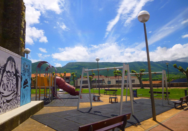 Spielplatz mit Schaukeln, Rutsche, Bänken, Masten, Lichtern, einer bemalten Wand, Gras, Bäumen, Gebäuden, Höhen und einem bewölktem Himmel im Hintergrund.