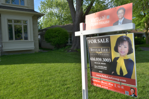 Ein Immobilien-Verkaufsschild steht vor einem Haus mit sichtbaren Fenstern, umgeben von grünem Gras, Bäumen und Pflanzen, unter einem Himmel im Hintergrund.