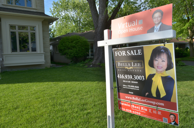 Ein Immobilien-Verkaufsschild steht vor einem Haus mit sichtbaren Fenstern, umgeben von grünem Gras, Bäumen und Pflanzen, unter einem Himmel im Hintergrund.
