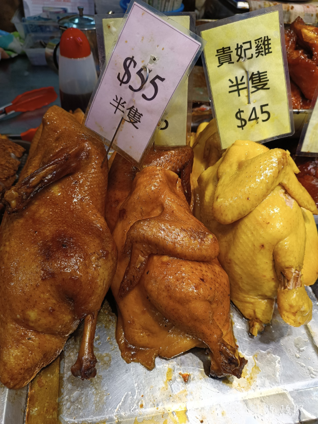 Eine Gruppe von Hühnern auf einem Metalltablett mit Preisschildern, vor einem Hintergrund verschiedener Marktartikel.