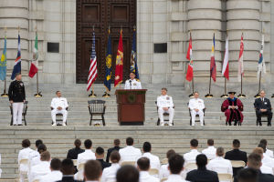 Gruppe von Menschen in weißen Uniformen auf Treppe sitzend mit einem Podium, das von Fahnen flankiert ist, vor einem großen Gebäude mit einer Tür.