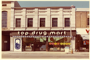 Eckladen "Top Drug Mart" mit Fenstern, einem Schild, einem Straßenpfosten, einem geparkten Fahrrad, einem Müllcontainer und einem bewölkten Himmel.