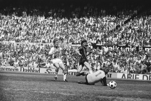 Schwarzes Foto von Männern, die auf einem Feld Füssball spielen, mit Zuschauern im Hintergrund und einem Banner "Bundesliga 1978-79 Ajax 1-1 Hannover 96".