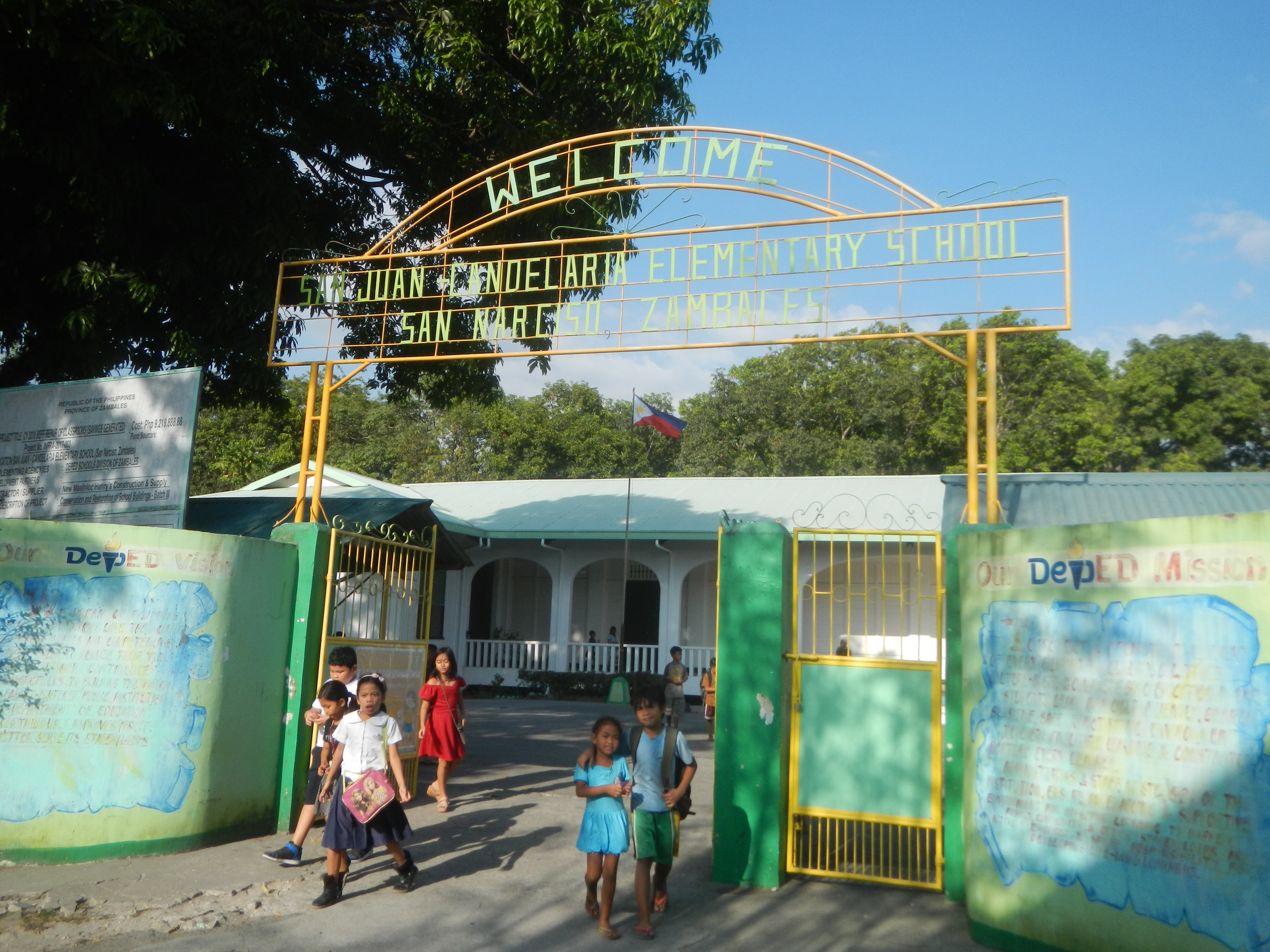 Eine Gruppe von Kindern, die vor einem Gebäude mit einem Torbogen gehen, umgeben von Bäumen und einem bewölkten Himmel.