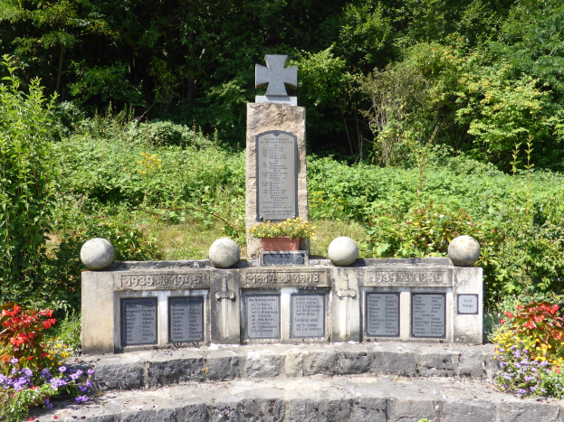 Grabstein mit Kreuz, beschriftet mit Text und Zahlen, in einem Garten umgeben von Pflanzen und Bäumen.