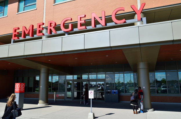 Gruppe von Menschen, die auf ein Notaufnahmelokal mit Glaswänden, Säulen, einem Namensschild, Schildern und einem Abfalleimer zugehen.