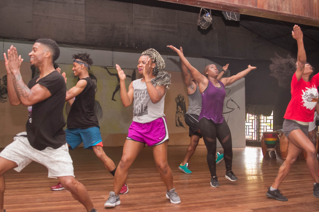 Eine Gruppe von Menschen, die in einem Studio mit einer bemalten Wand, einem Fenster und Deckenbeleuchtung tanzen, was auf einen Zumba-Kurs hinweist.