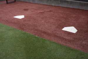 Baseballfeld mit Kunstrasen, umgeben von einem Zaun, mit Home Plate in der Mitte und einer Wand im Hintergrund.