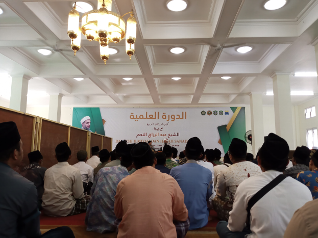 Eine Gruppe von Menschen, die auf dem Boden vor einer Fahne mit Text und einem Bild einer Person sitzen, umgeben von Säulen und beleuchtet von Lampen und Kronleuchtern, mit einem Fenster auf der rechten Seite.