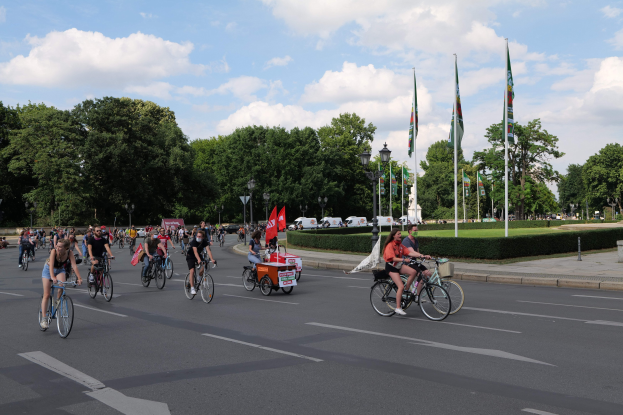 Eine Gruppe von Radfahrern fährt eine von Bäumen gesäumte Straße mit Laternenpfählen und Fahnenmasten entlang, mit Fahrzeugen und bewölktem Himmel im Hintergrund während der Paris Bike Ride 2016.