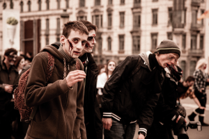 Eine Gruppe von Menschen in Zombie-Kostümen, einige in Verkleidung, gehen eine Straße entlang mit einem Gebäude im Hintergrund, eine Person hält eine Kamera.