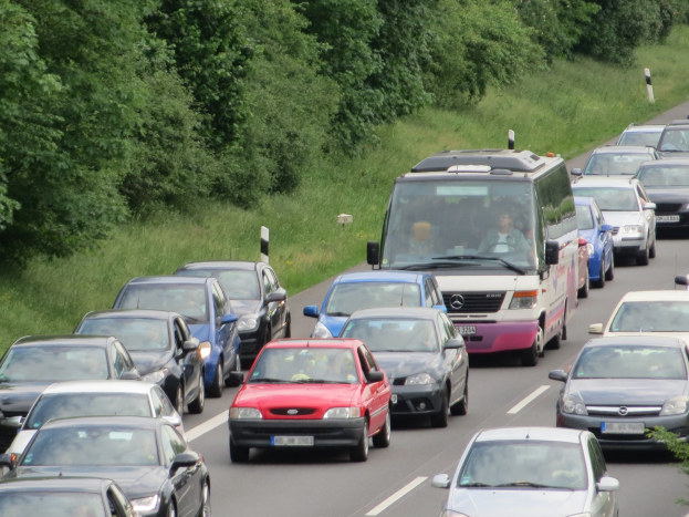 Verkehrsstau auf einer Autobahn mit mehreren Autos und einem Van, Menschen sichtbar in den Fahrzeugen und Bäume und Gras im Hintergrund.