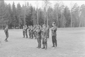 Schwarz-weißes Bild einer Gruppe von Männern in Mützen, die Gewehre während eines Trainingsübungs in einem offenen Feld mit Bäumen und einem klaren Himmel halten.