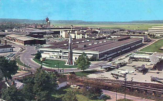 Alte Schwarz-Weiß-Aufnahme eines Flughafens mit geparkten Propellerflugzeugen, Terminalgebäuden, Bäumen, Straßen, Autos, Strommasten, Gras, Bahnschienen, fernen Türmen, Hügeln und einem klaren Himmel, mit gedrucktem Text am unteren Rand.