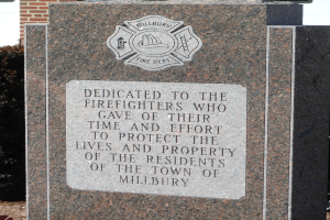 Denkmalstein mit einer Gedenktafel für Feuerwehrleute in Millbury, vor einem Backsteingebäude mit umliegender Vegetation gesetzt.