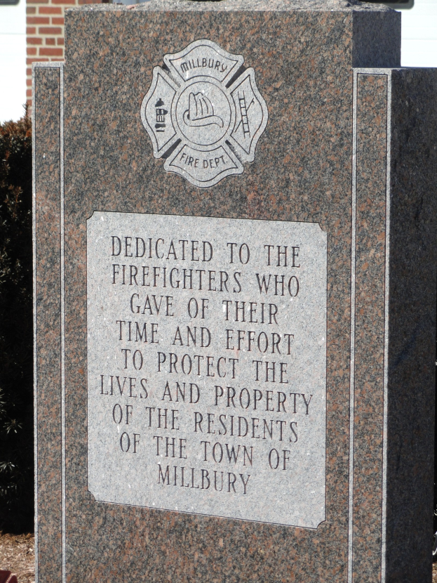 Denkmalstein mit einer Gedenktafel für Feuerwehrleute in Millbury, vor einem Backsteingebäude mit umliegender Vegetation gesetzt.