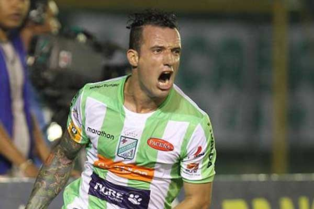 Ein Fußballspieler in grüner und weißer Uniform mit sichtbaren Tattoos auf dem Arm feiert ein Tor, mit unscharfem Hintergrund und einer Tafel.