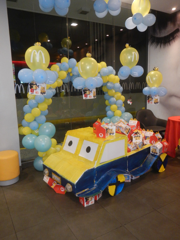 Eine McDonald's-Geburtstagsparty-Szene mit einem gelben und blauen Lastwagen, der mit Ballons und anderen Dekorationen geschmückt ist, einem rot eingedeckten Tisch mit Gegenständen, Hockern, einer Glaswand und Deckenlampen, die eine festliche Atmosphäre schaffen.
