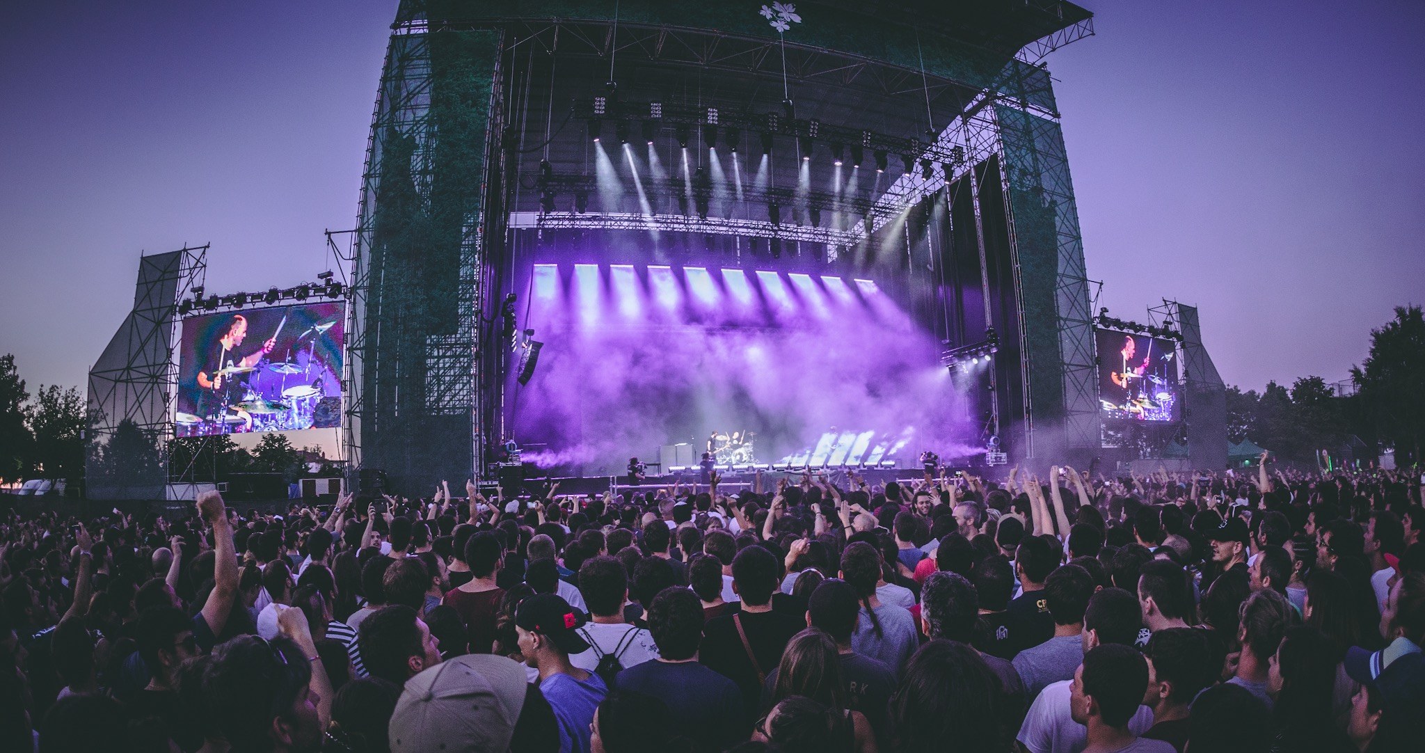 Eine große Menge bei einem Musikfestival schaut auf der hell erleuchteten Bühne mit Instrumenten und Bildschirmen, die vor einem Gebäude, Bäumen und einem klaren blauen Himmel liegt.