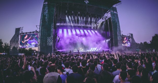 Eine große Menge bei einem Musikfestival schaut auf der hell erleuchteten Bühne mit Instrumenten und Bildschirmen, die vor einem Gebäude, Bäumen und einem klaren blauen Himmel liegt.