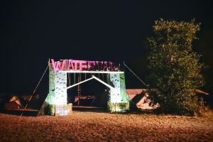 Bühne für ein nächtliches Musikfestival mit einem Bogen mit Text, einem Baum auf der rechten Seite, Zelten im Hintergrund und einem dunklen Himmel.