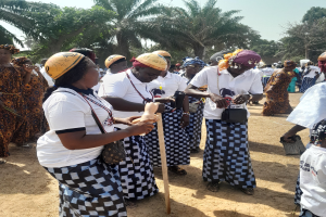 Gruppe von Frauen in weißen und schwarzen Kleidern mit einigen, die gelbe Helme tragen, auf einem Feld mit Bäumen und einem klaren blauen Himmel im Hintergrund.