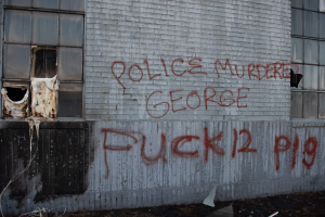 Graffiti auf einer Hauswand mit der Aufschrift "Polizei ermordet George Fuck 12 P19" mit Fenstern und verstreuten Gegenständen davor.