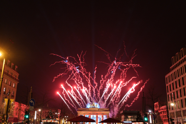 Eine belebte Stadtstraße am Neujahrstag in Berlin, voller Menschen, Fahrzeugen und Gebäuden, erhellt von Lichtern und Feuerwerk am Himmel.