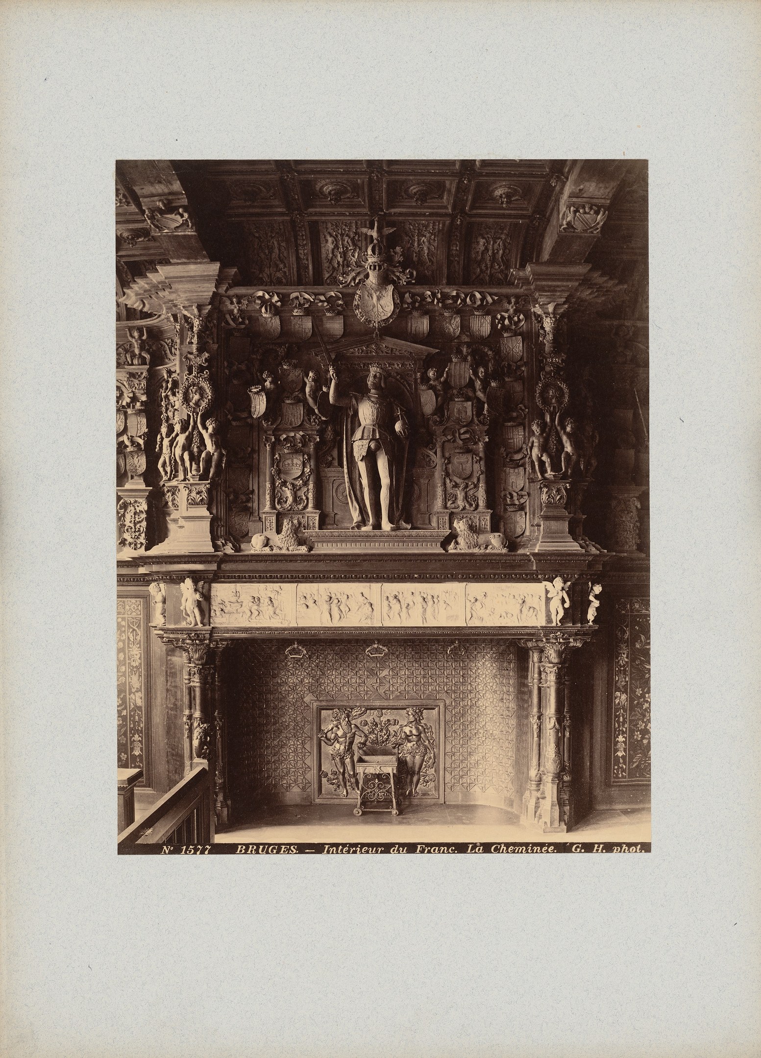 Altes Schwarz-Weiß-Foto eines Kamins mit einer zentralen Statue, umgeben von Wandskulpturen und einer dekorierten Decke, mit Text am unteren Rand.